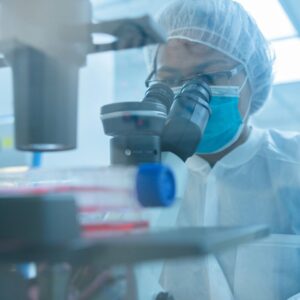 Umbilical Cord Stem Cells for Heart Failure A scientist wearing a lab coat, face mask, and hairnet looks through a microscope in a blue-toned laboratory setting, with a cell culture flask visible in the blurred foreground.